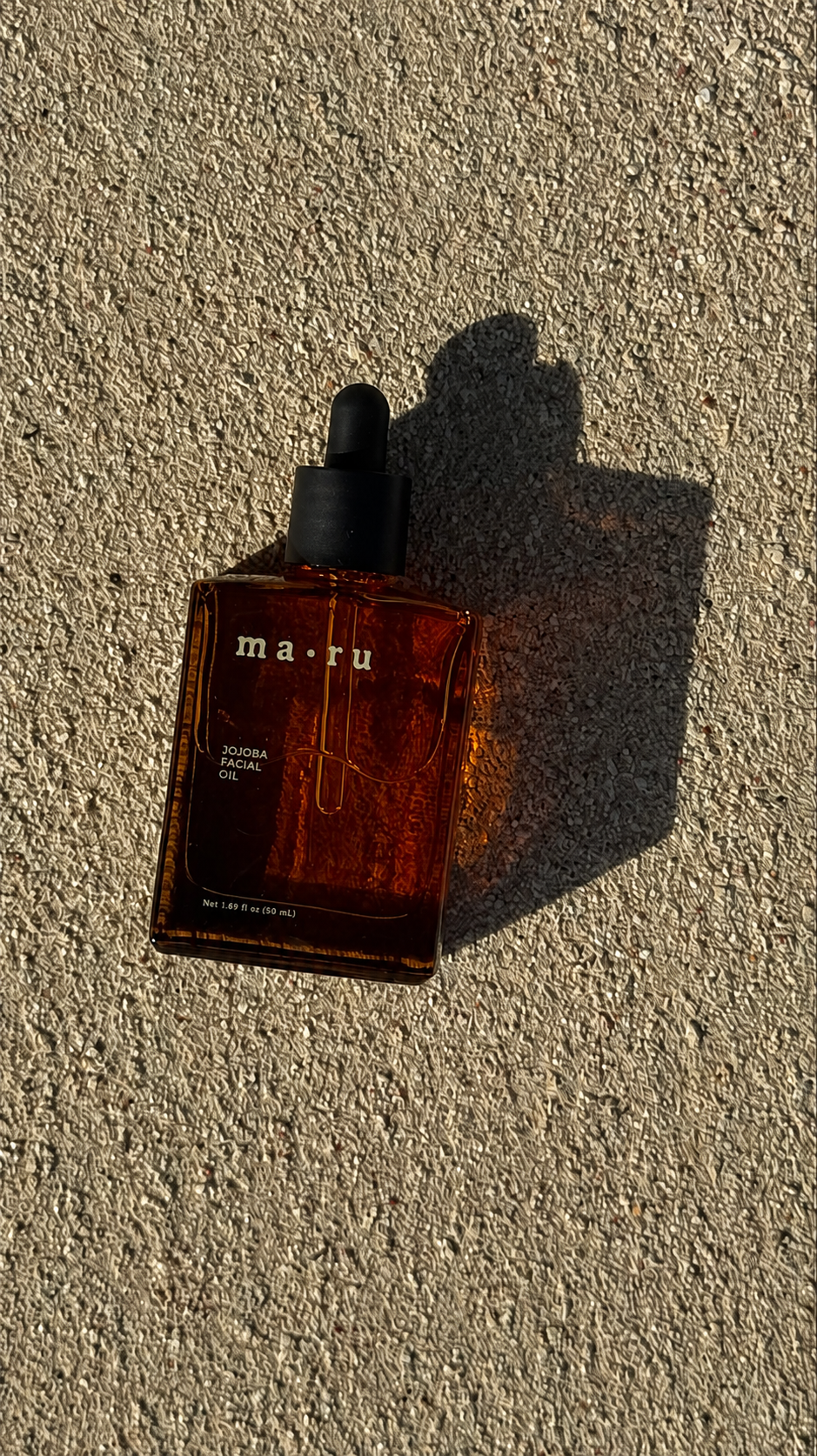 A square amber glass dropper bottle of MARU Jojoba Facial Oil, labeled “ma·ru” with a black rubber dropper cap, placed on a textured concrete surface in direct sunlight casting a strong shadow, highlighting a minimalist botanical face oil designed to hydrate, nourish and support soft, smooth, radiant-looking skin.