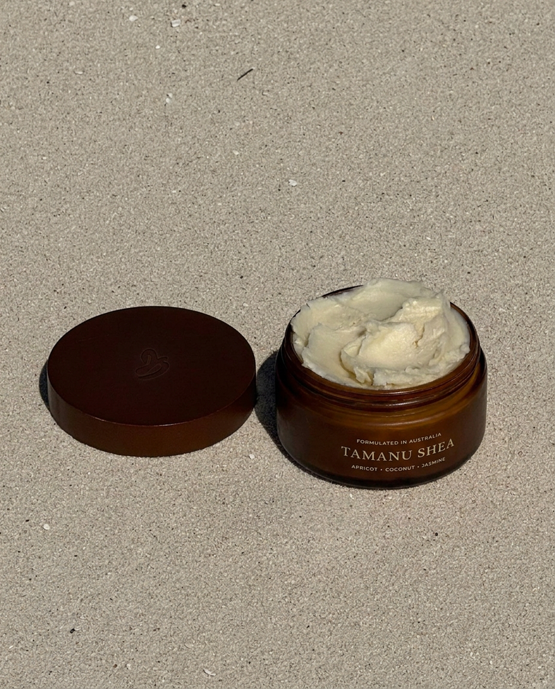 An open amber jar of Tamanu Shea body butter with its lid placed beside it on a sandy surface. The jar contains a thick, pale cream-colored product, and the label reads 'TAMANU SHEA' with 'JASMINE + COCONUT + APRICOT underneath.