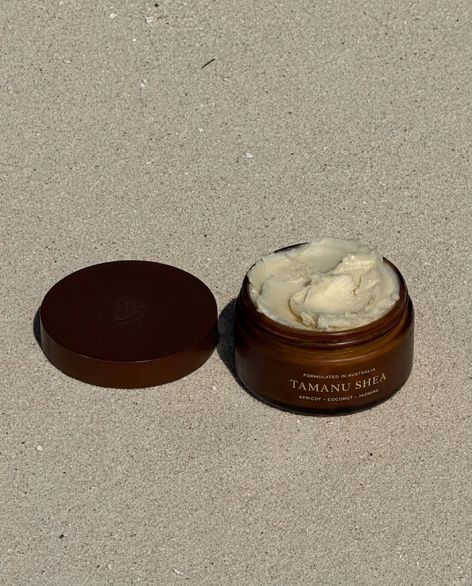 An open amber jar of Tamanu Shea body butter with its lid placed beside it on a sandy surface. The jar contains a thick, pale cream-colored product, and the label reads 'TAMANU SHEA' with 'JASMINE + COCONUT + APRICOT underneath.