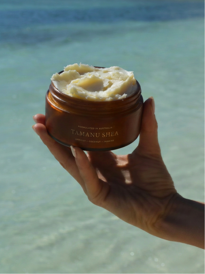 A hand holding an open amber jar of Tamanu Shea hand and body butter, with a blurred beach and turquoise ocean background.